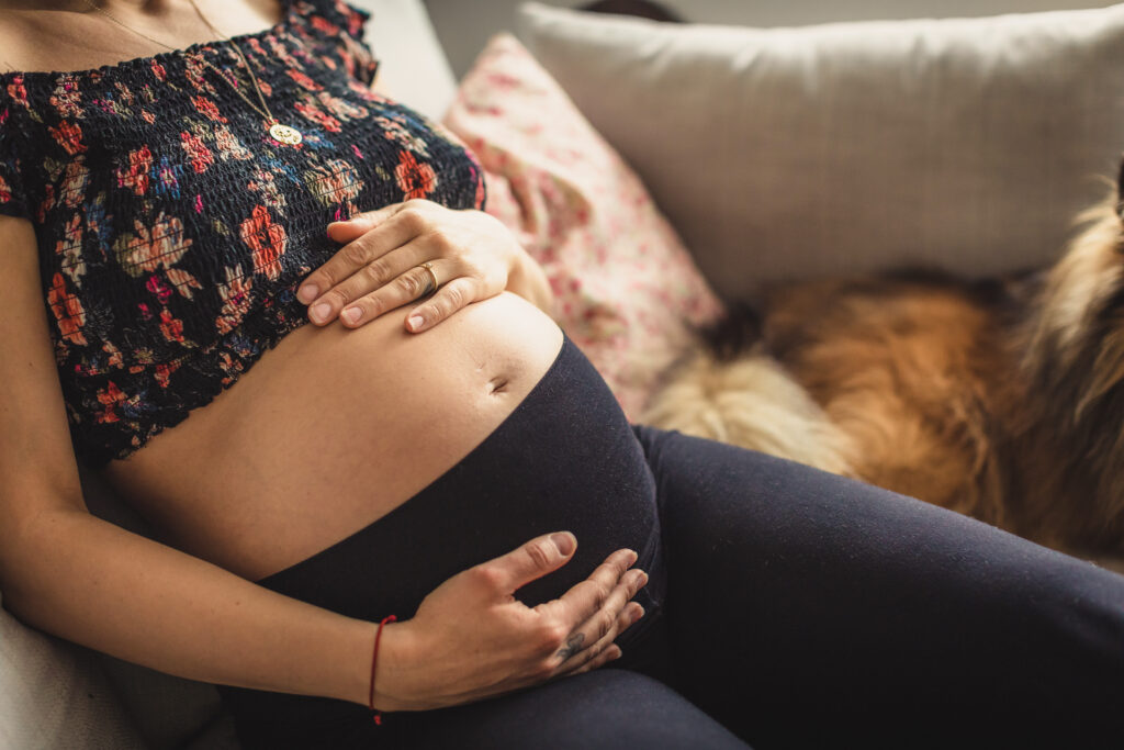 Babybauchfotos Bremen – Frau streichelt Bauch auf Sofa mit Hund im Hintergrund